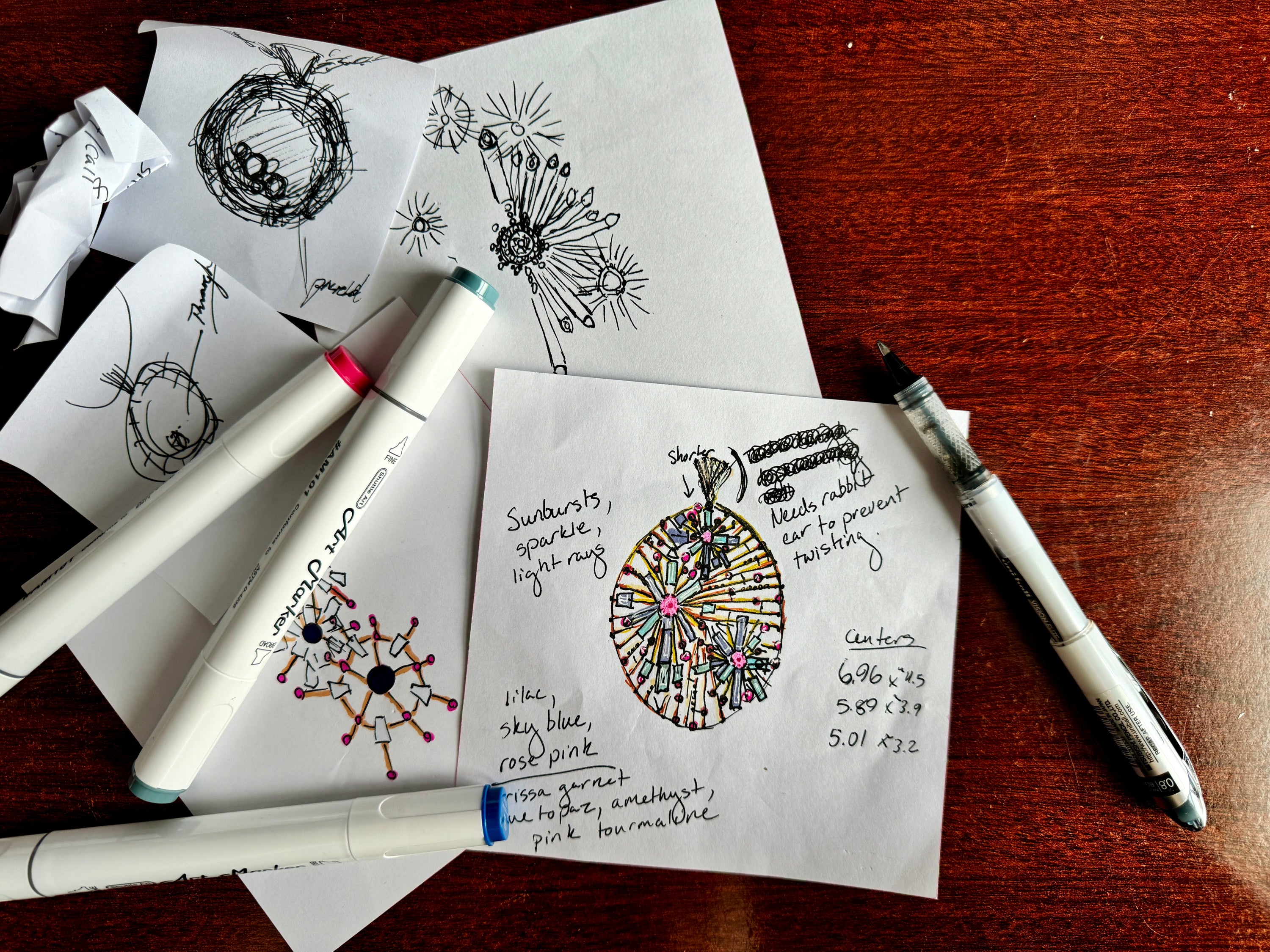 rough jewelry sketches on paper with markers on a wooden surface